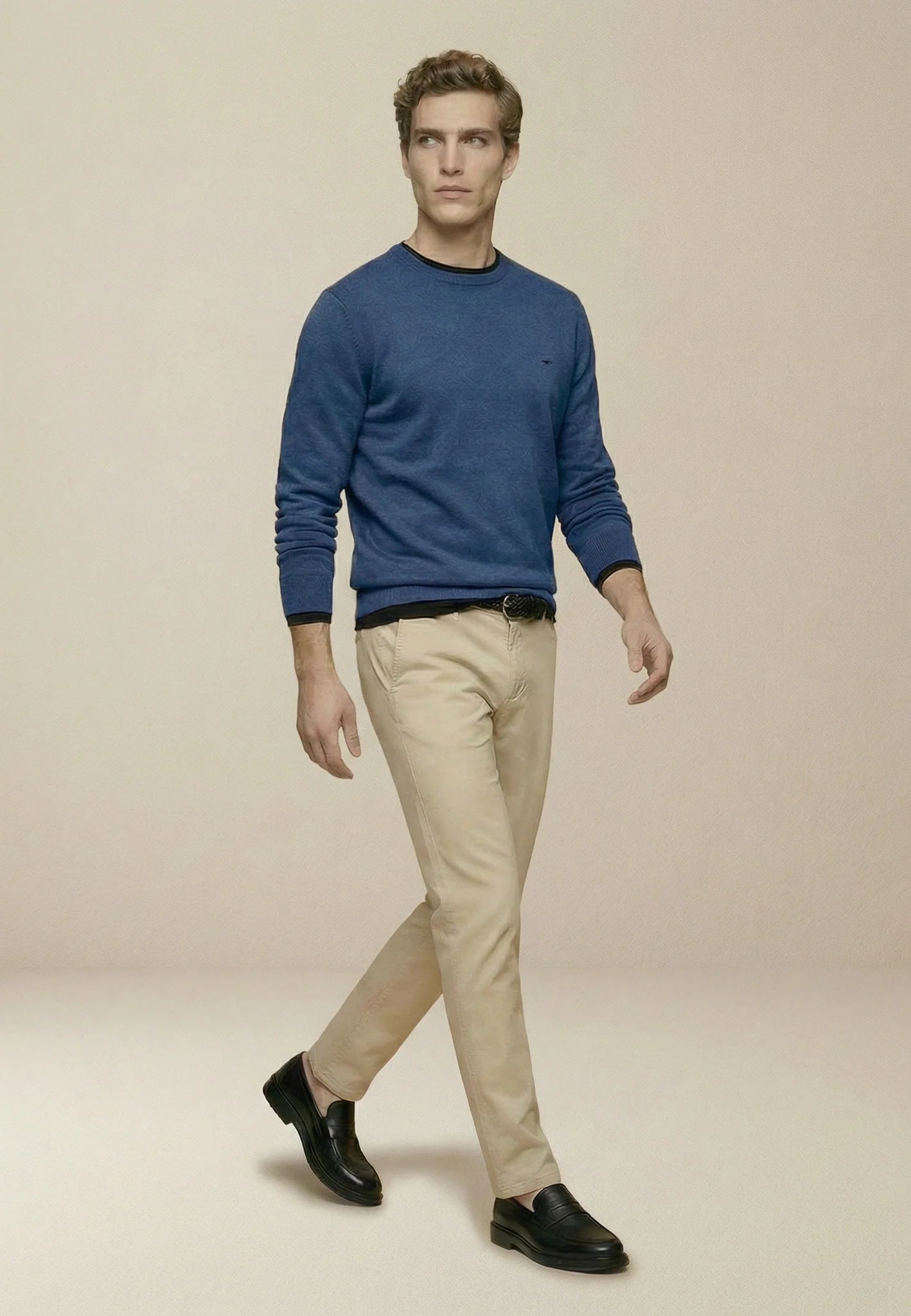 Man wearing a blue sweater, beige pants, and black shoes on a beige background