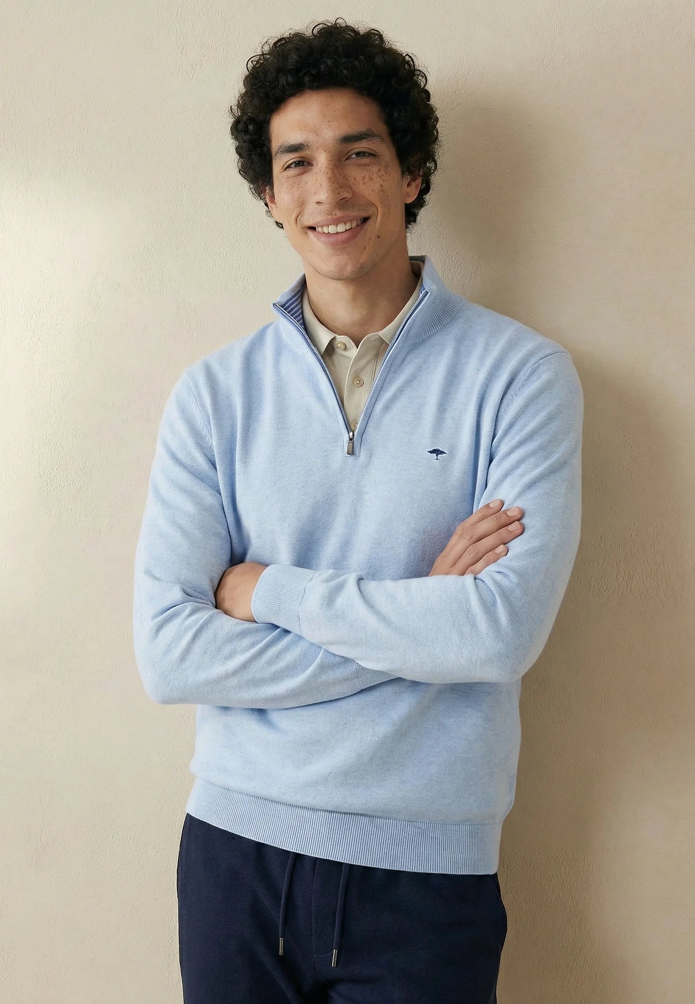 Man wearing a light blue sweater with a brand logo, standing against a beige wall.