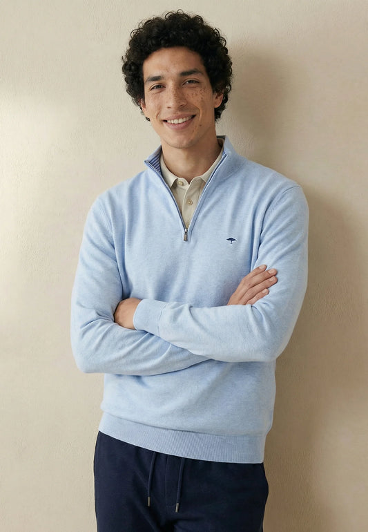 Man wearing a light blue sweater with a brand logo, standing against a beige wall.