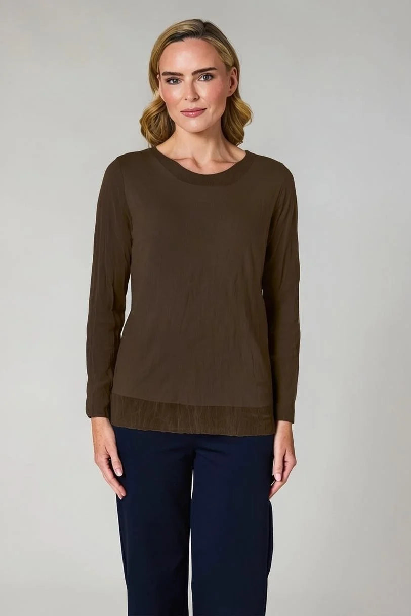 A woman modeling a dark brown long-sleeve knit top with a crew neck and a subtle textured hem, paired with navy blue trousers, against a neutral light gray background.