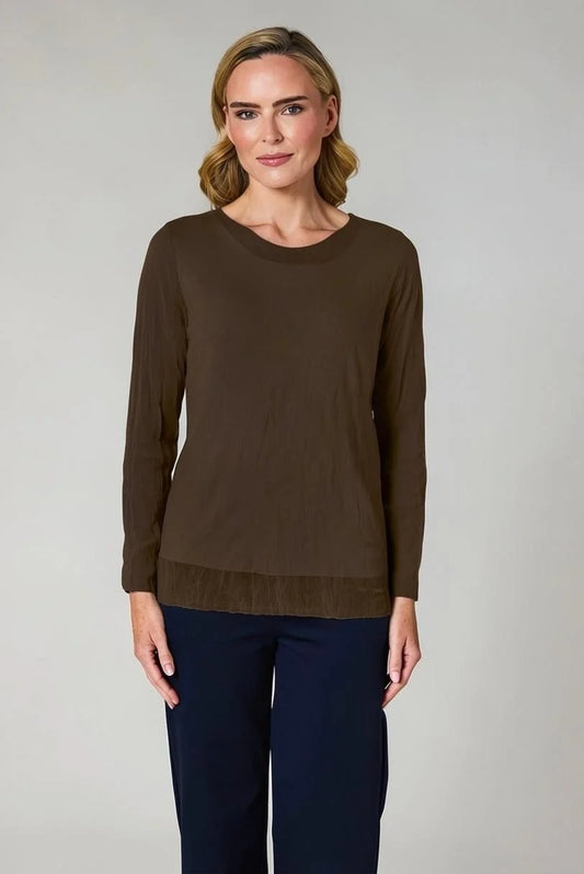 A woman modeling a dark brown long-sleeve knit top with a crew neck and a subtle textured hem, paired with navy blue trousers, against a neutral light gray background.