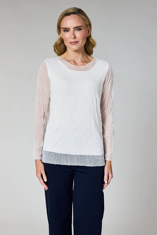 A woman modeling a white long-sleeve top with sheer blush-beige mesh sleeves and a sheer gray textured hem, paired with navy trousers, against a neutral gray studio background.