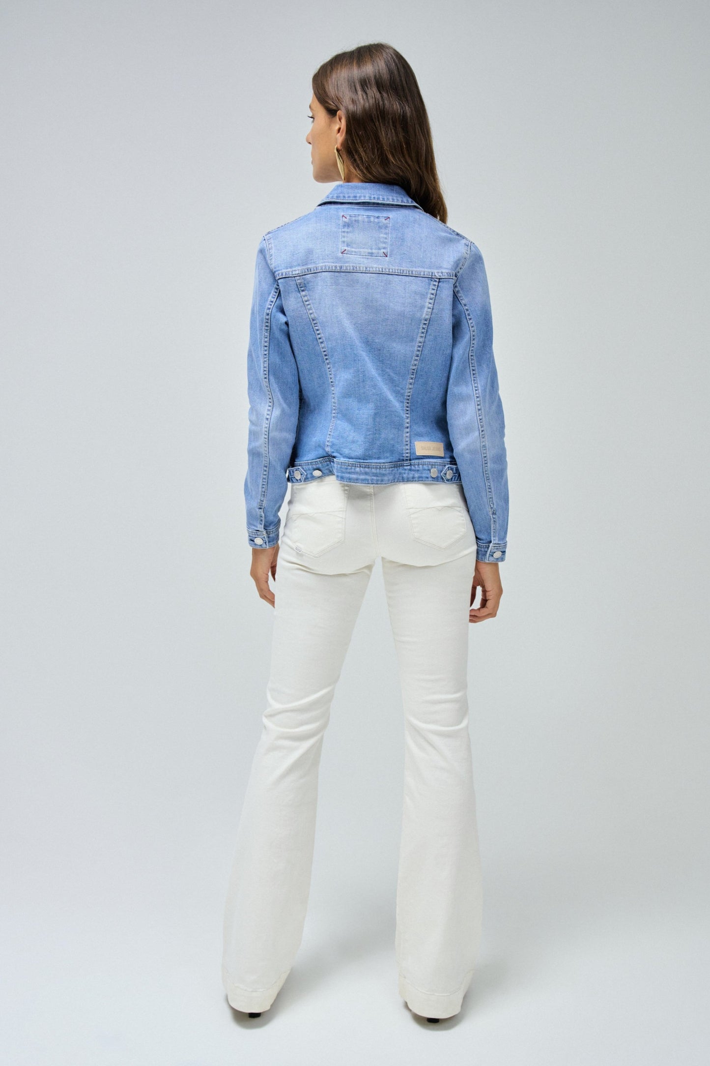 Woman wearing a light blue denim jacket and white pants on a gray background