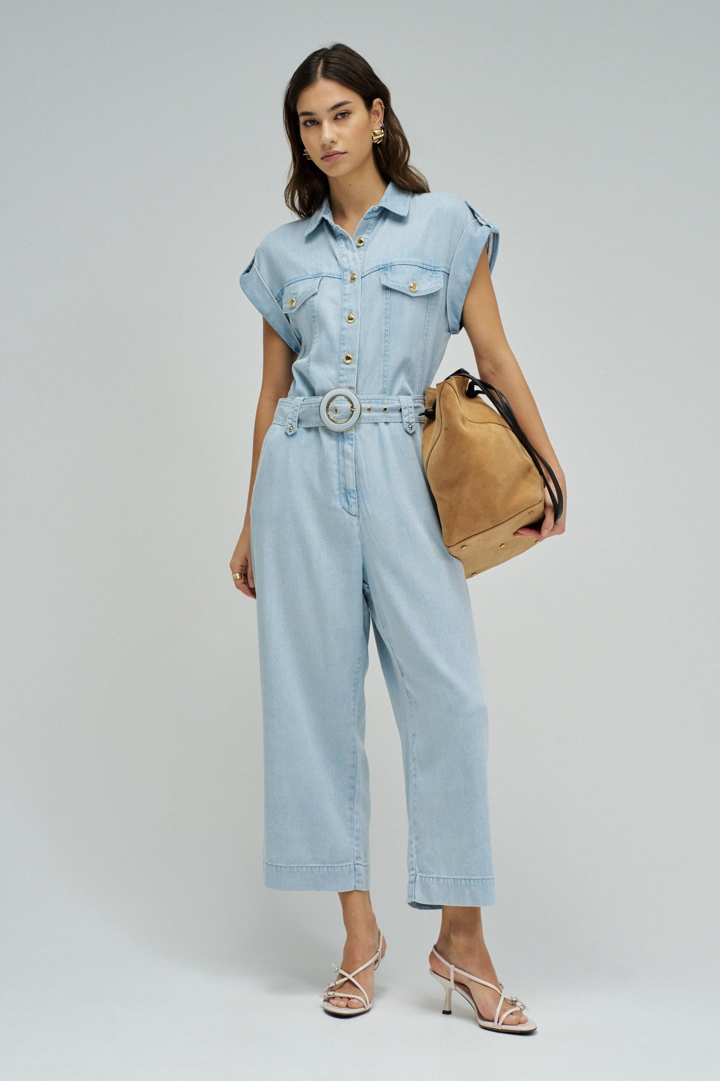 Woman wearing a light blue denim jumpsuit with a beige bag on a white background