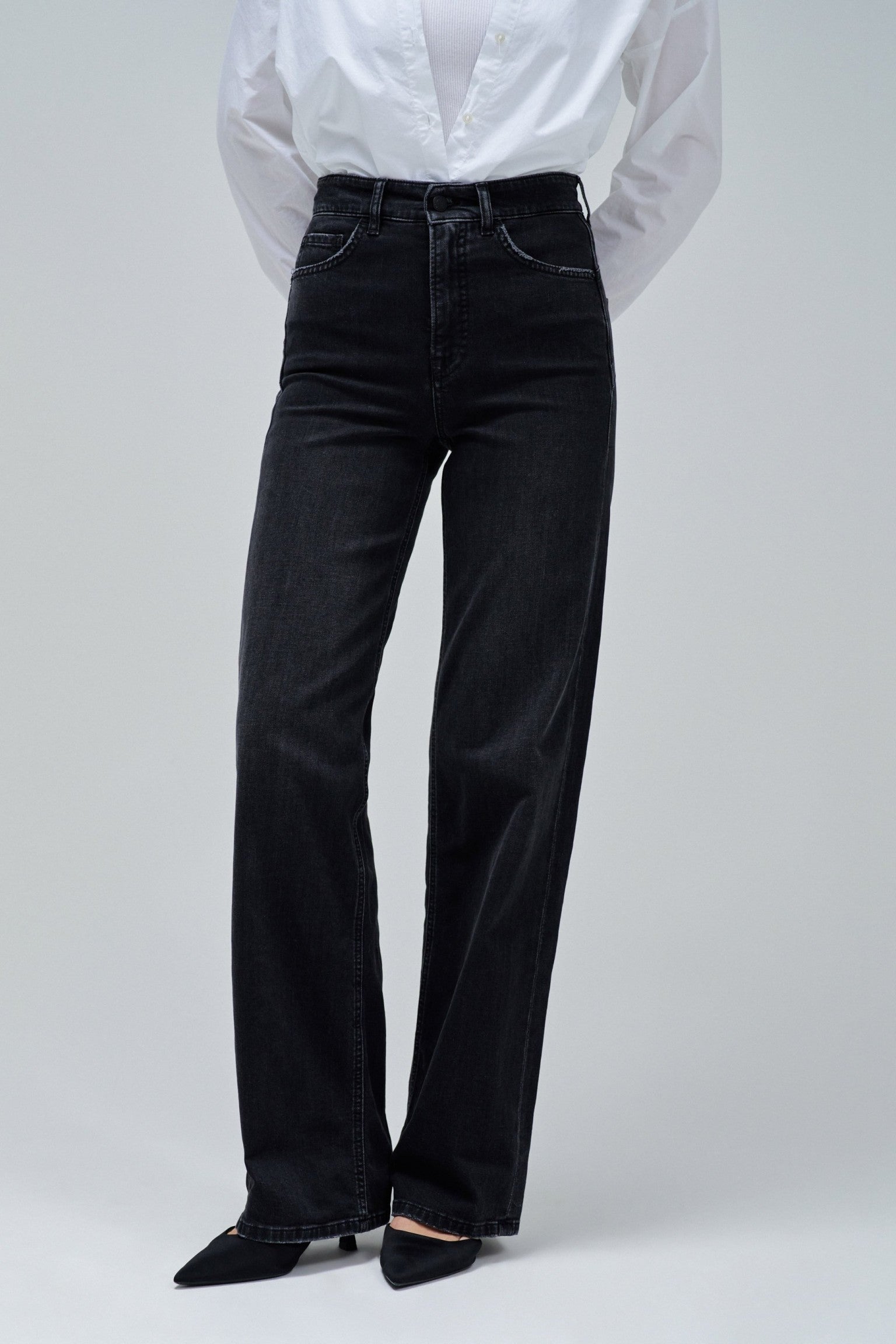 A person wearing black wide-leg jeans with a white button-up shirt tucked in and black pointed shoes, photographed against a light gray studio background.