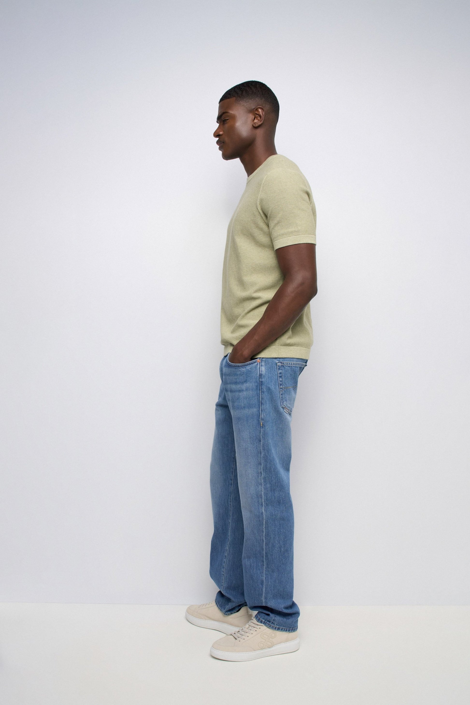 Man wearing a light green t-shirt and blue jeans standing against a white background