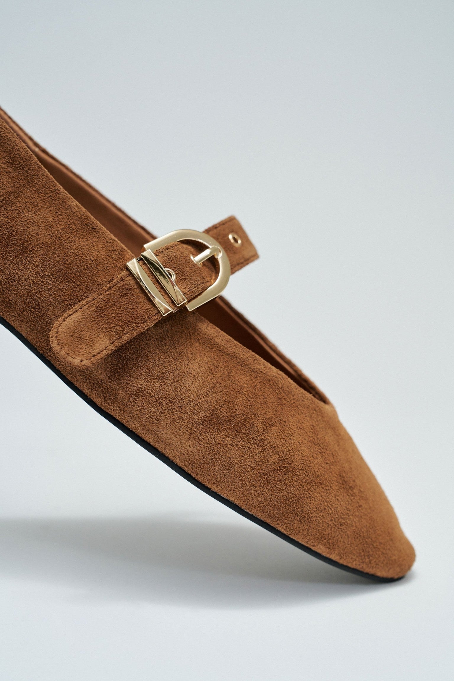 A close-up of a brown suede loafer with a pointed toe and a gold-tone buckle detail on a light gray background.