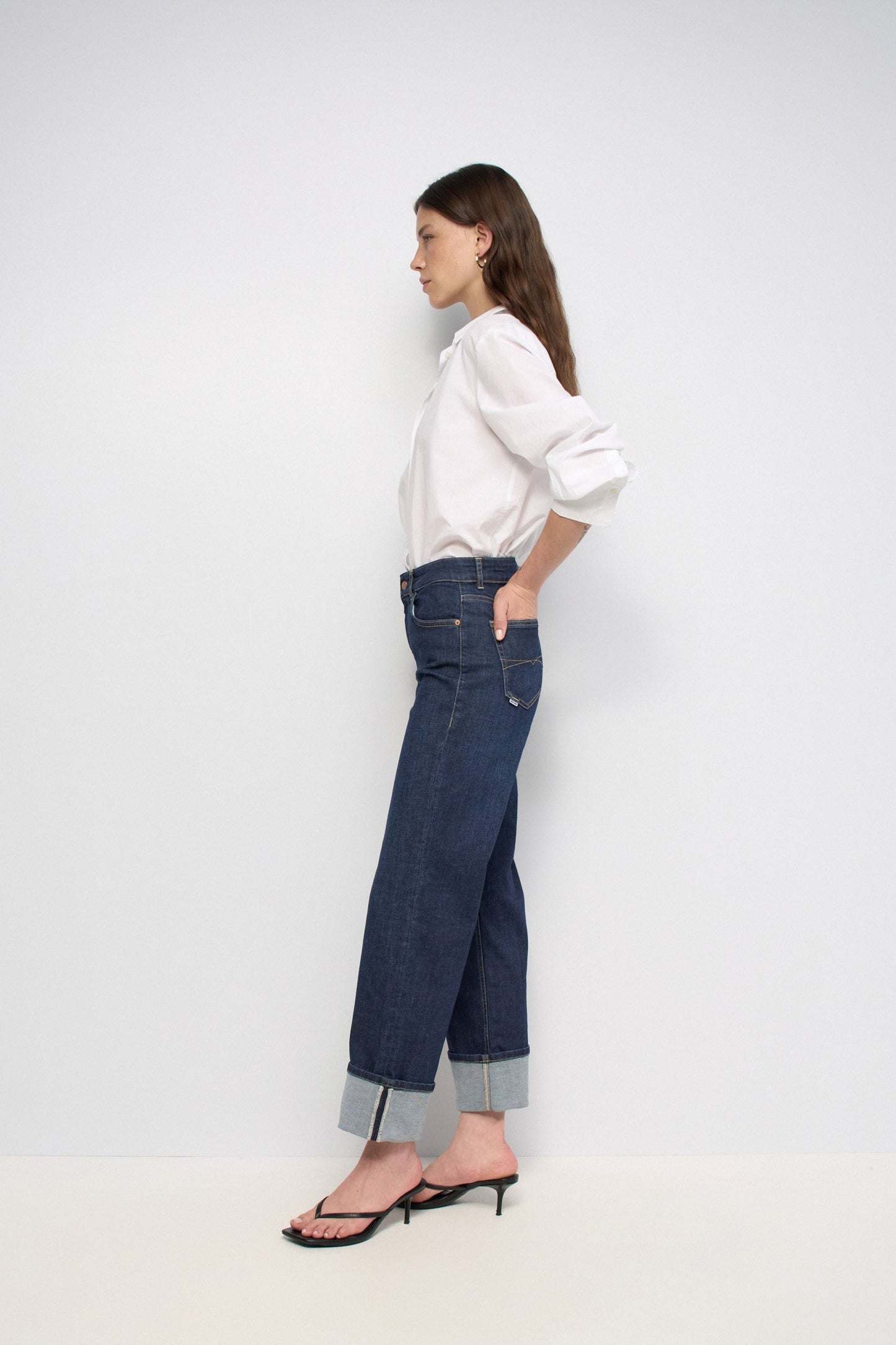 A woman stands in profile wearing dark blue wide-leg cuffed jeans, a white long-sleeve blouse, and black heeled sandals against a plain light gray studio background.