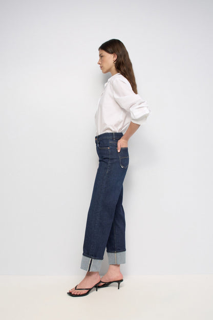 A woman stands in profile wearing dark blue wide-leg cuffed jeans, a white long-sleeve blouse, and black heeled sandals against a plain light gray studio background.