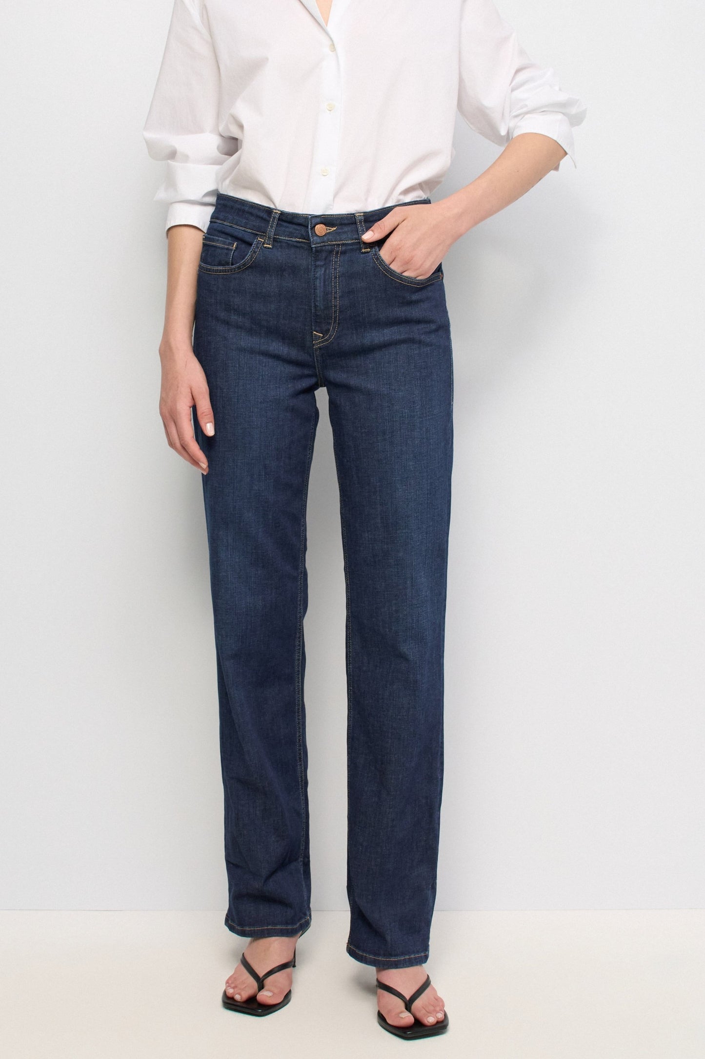 A model wearing dark blue straight-leg denim jeans, a white button-down shirt, and black sandals, standing against a plain white studio background.
