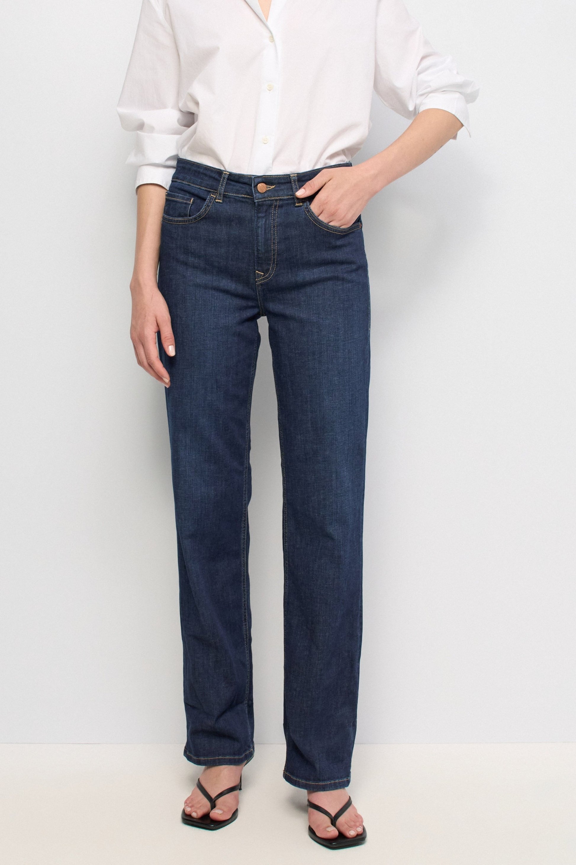 A model wearing dark blue straight-leg denim jeans, a white button-down shirt, and black sandals, standing against a plain white studio background.