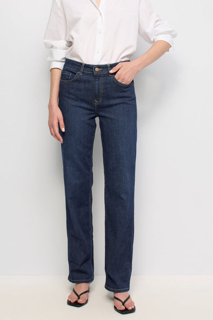 A model wearing dark blue straight-leg denim jeans, a white button-down shirt, and black sandals, standing against a plain white studio background.