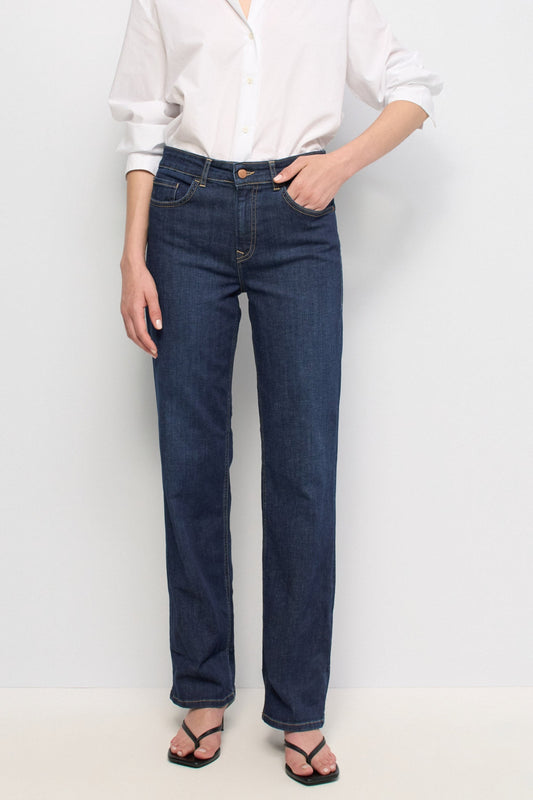 A model wearing dark blue straight-leg denim jeans, a white button-down shirt, and black sandals, standing against a plain white studio background.
