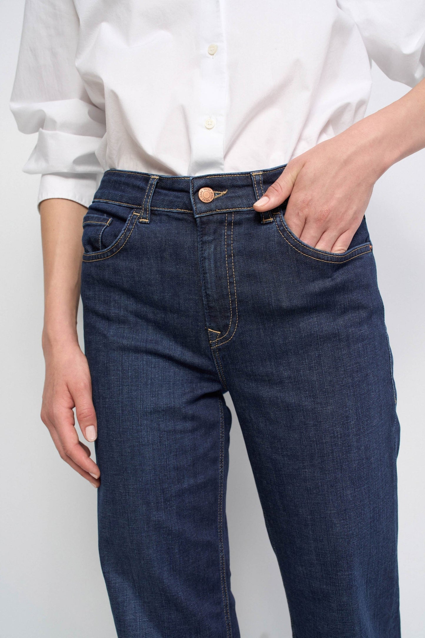 A close-up of a person wearing dark blue denim jeans with a white button-up shirt tucked in, standing against a light gray studio background.