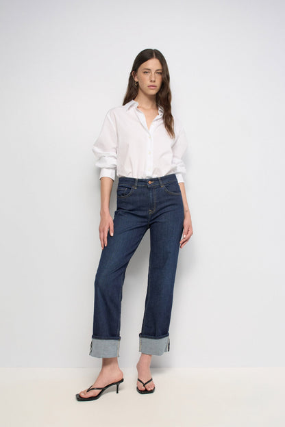 A woman modeling a pair of dark blue straight-leg jeans with rolled cuffs, paired with a white button-down shirt and black heeled sandals, against a plain white studio background.