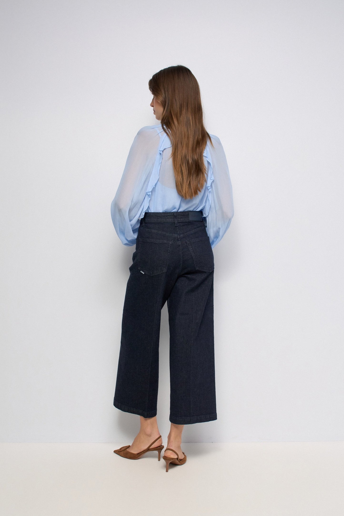 A woman is shown from behind wearing dark wide-leg cropped jeans, a light blue sheer long-sleeve blouse, and brown slingback heels against a plain light gray studio background.