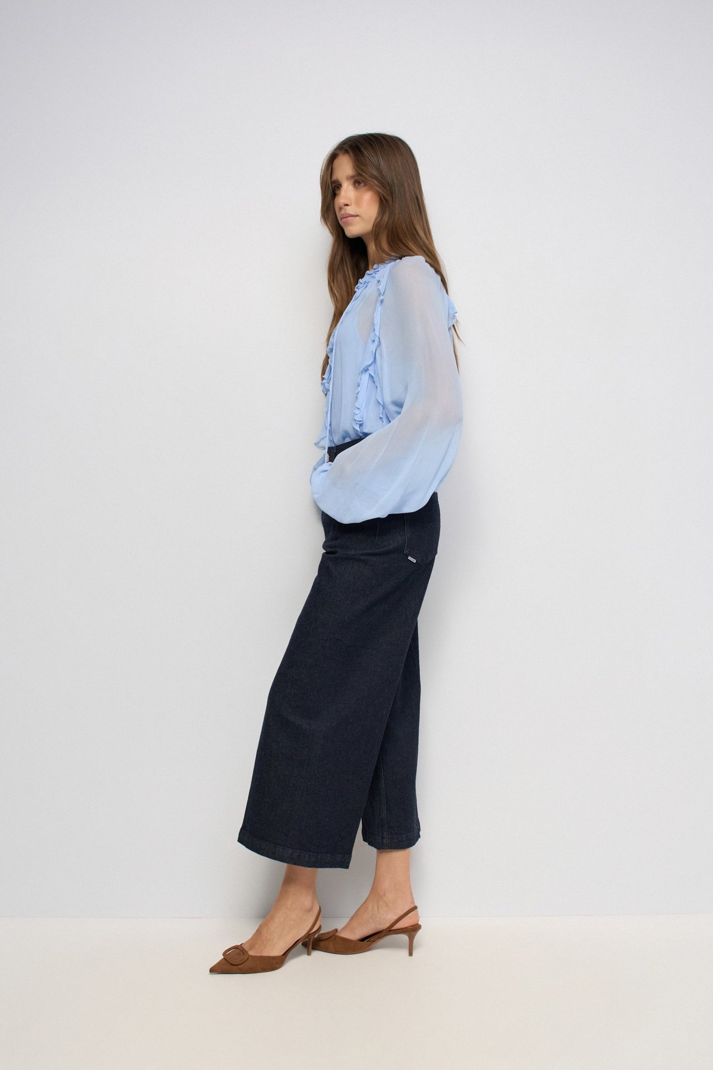 A woman posing in a studio wearing a light blue sheer ruffled blouse, dark navy cropped wide-leg jeans, and brown slingback heels against a plain light gray background.