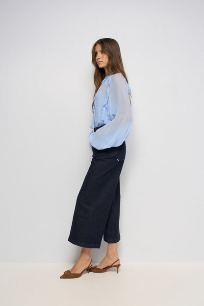 A woman posing in a studio wearing a light blue sheer ruffled blouse, dark navy cropped wide-leg jeans, and brown slingback heels against a plain light gray background.