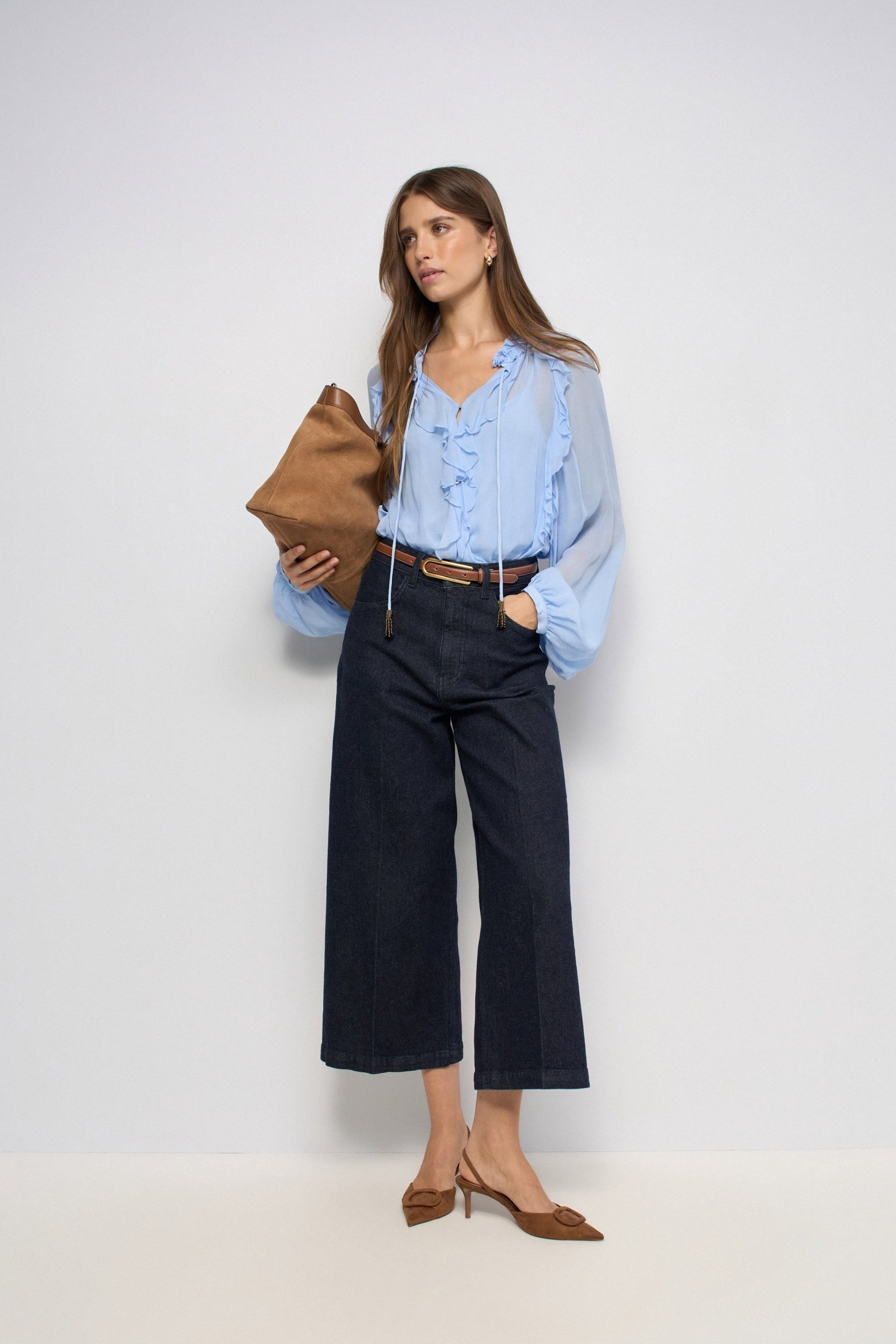 A woman modeling a light blue ruffled blouse, dark wide-leg denim pants, and brown pointed-toe heels, holding a brown clutch against a plain light gray studio background.