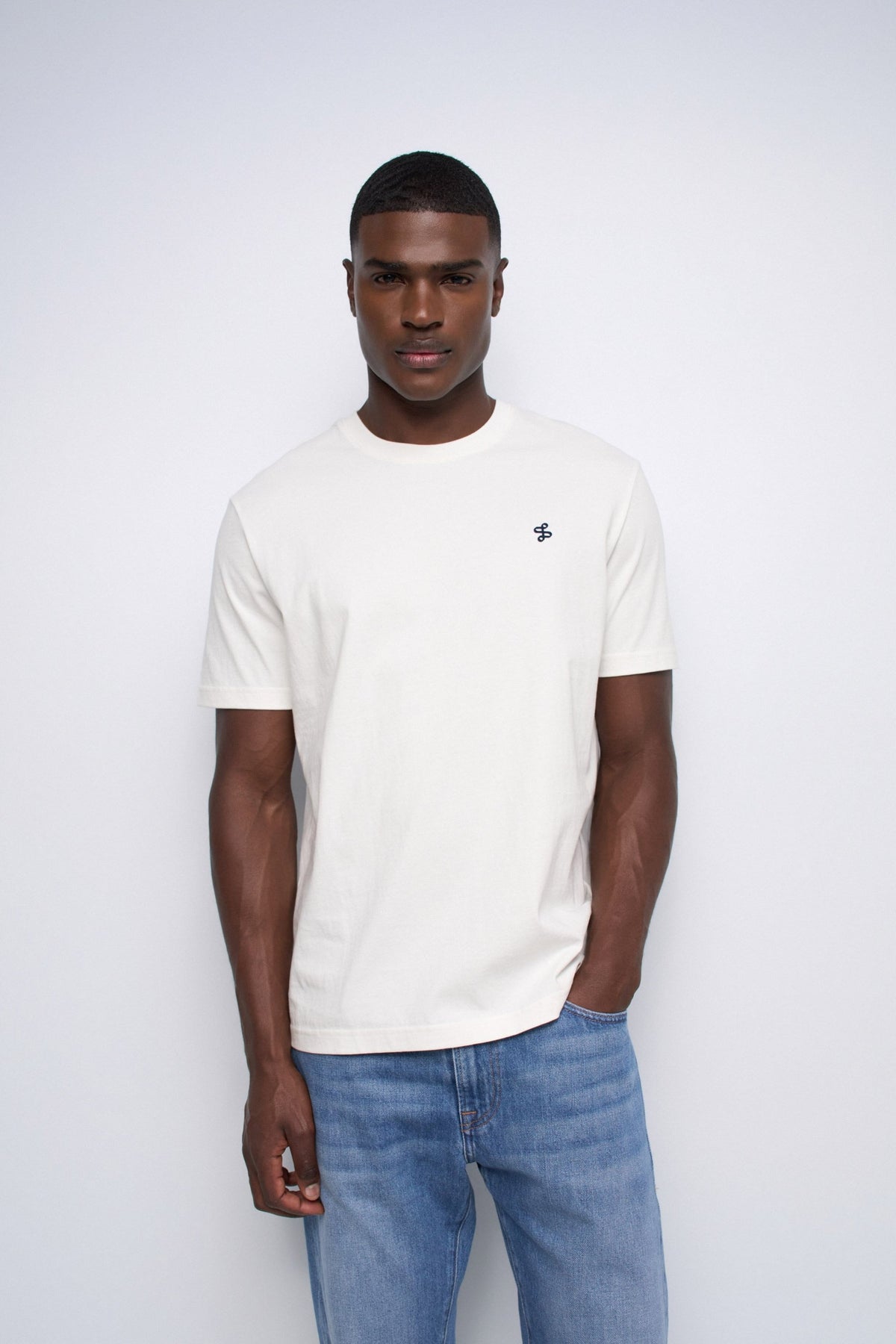 A man wearing a cream-colored short-sleeve crewneck t-shirt with a small embroidered logo on the left chest, paired with blue jeans, standing against a light gray studio background.