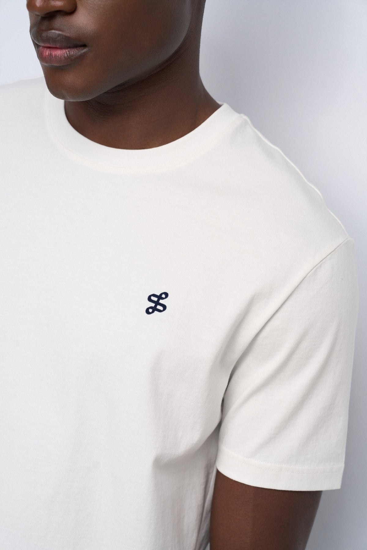 Close-up of a person wearing a white short-sleeve crewneck t-shirt with a small black embroidered logo on the left chest, set against a light gray background.