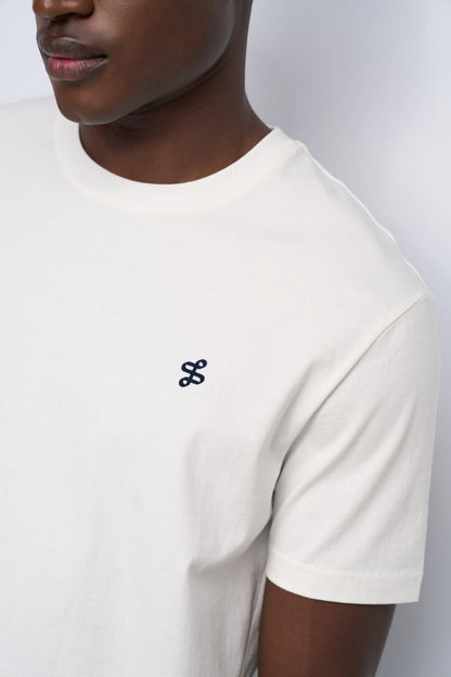 Close-up of a person wearing a white short-sleeve crewneck t-shirt with a small black embroidered logo on the left chest, set against a light gray background.