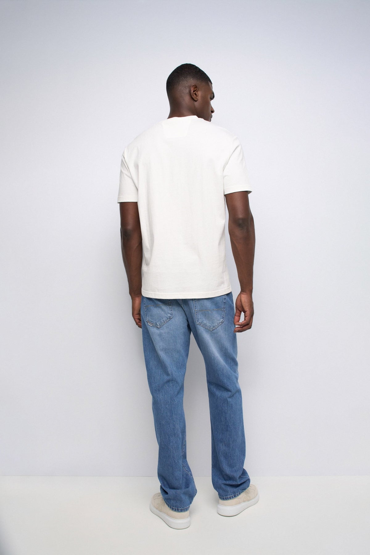A man is shown from behind wearing a white crew-neck t-shirt, blue jeans, and white sneakers against a plain white studio background.