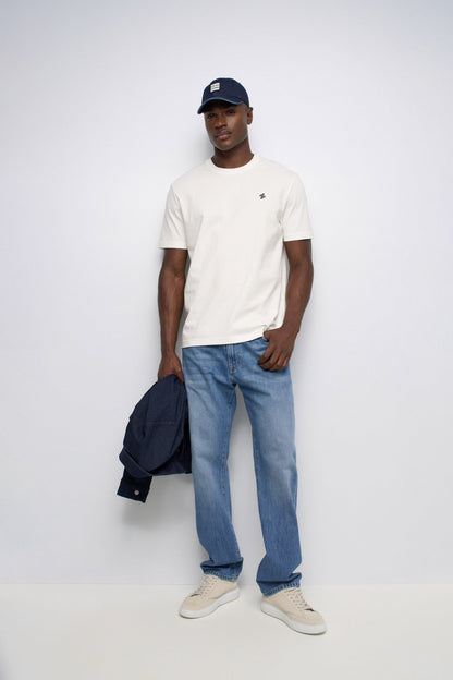 A man modeling a white crew-neck t-shirt with a small embroidered logo on the chest, paired with blue jeans, a navy blue cap, and holding a navy jacket, against a plain light gray studio background.