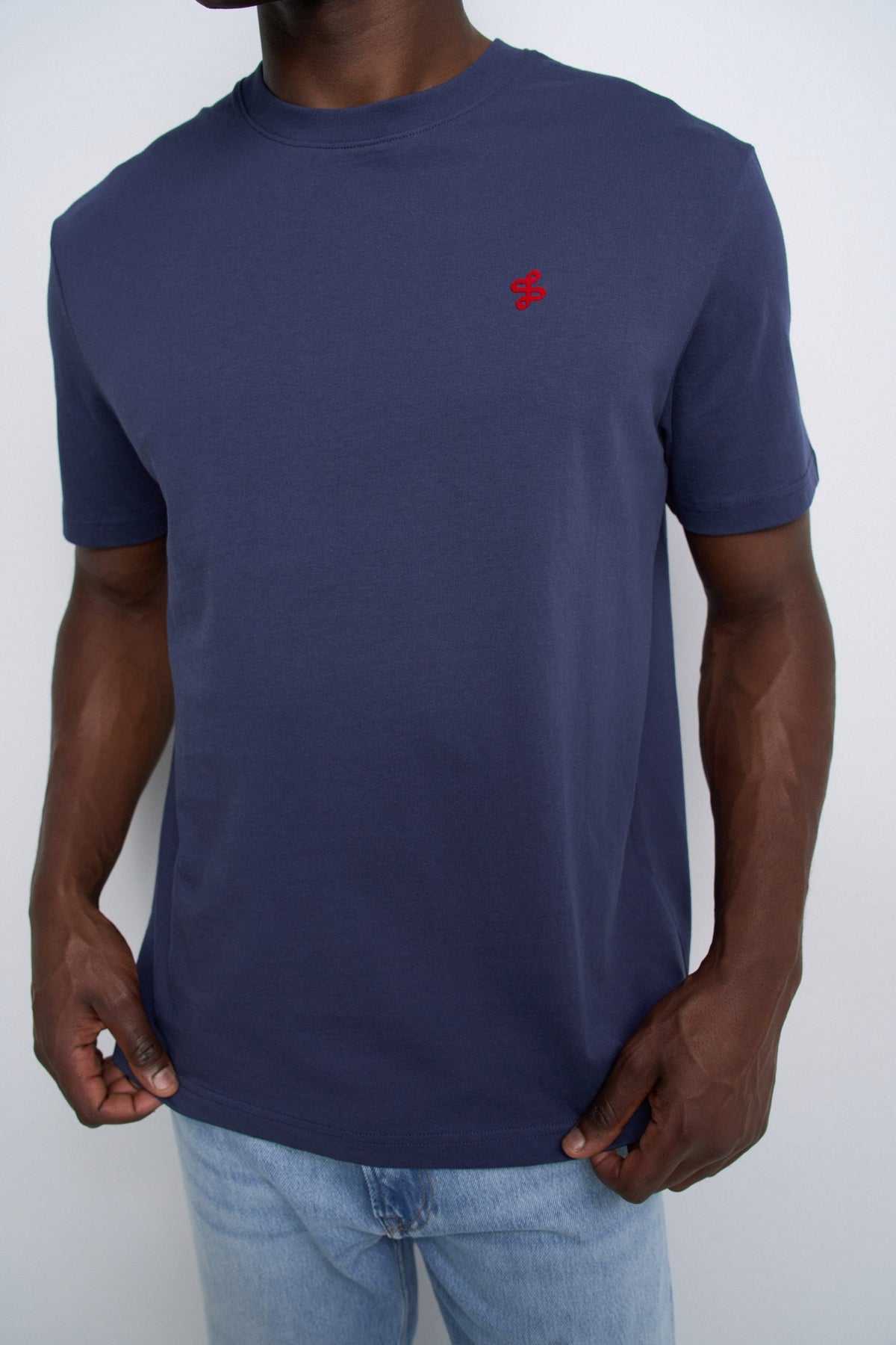 A man wearing a navy blue short-sleeve crewneck t-shirt with a small red embroidered logo on the left chest, paired with light blue jeans.