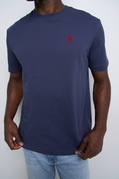 A man wearing a navy blue short-sleeve crewneck t-shirt with a small red embroidered logo on the left chest, paired with light blue jeans.