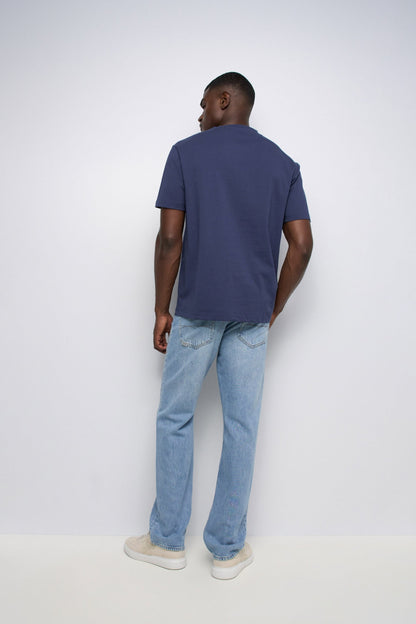 A man viewed from behind wearing a navy blue short-sleeve t-shirt, light blue jeans, and off-white sneakers, standing against a plain light gray studio background.