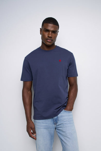 A man wearing a navy blue crewneck t-shirt with a small red embroidered logo on the chest, paired with light blue jeans, standing against a light gray studio background.