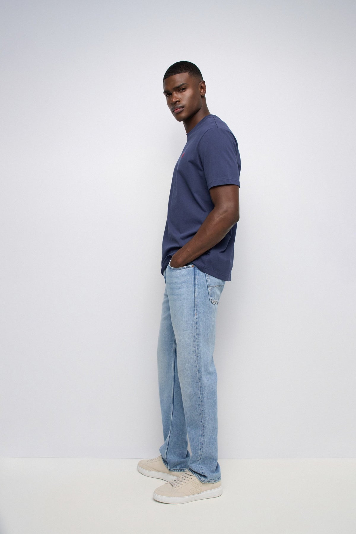 A man wearing a navy blue crewneck t-shirt, light wash blue jeans, and beige sneakers, standing in profile against a plain white studio background.