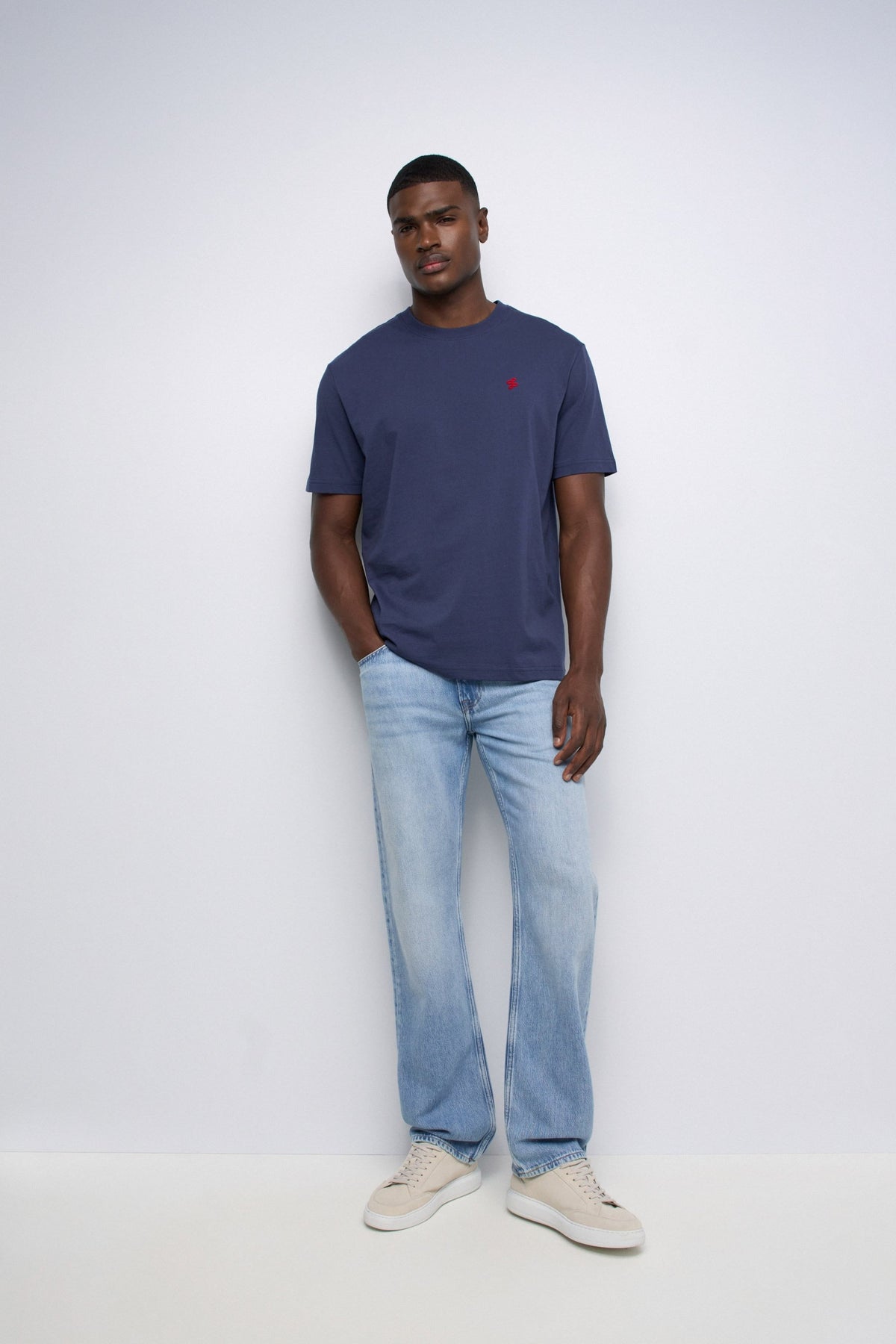 A man wearing a navy blue crew-neck t-shirt with a small red embroidered logo on the chest, paired with light-wash blue jeans and beige sneakers, standing against a plain white studio background.