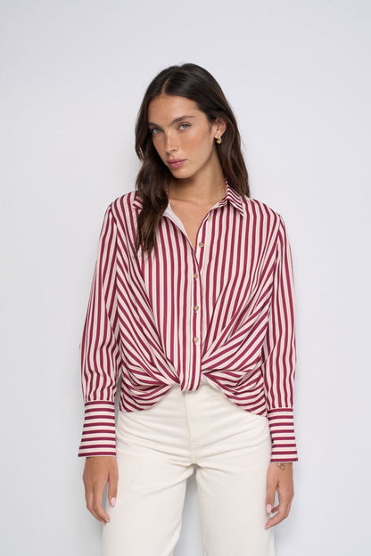 A woman modeling a red and white vertically striped button-down shirt with a twisted knot at the waist, paired with off-white pants, against a plain light gray background.