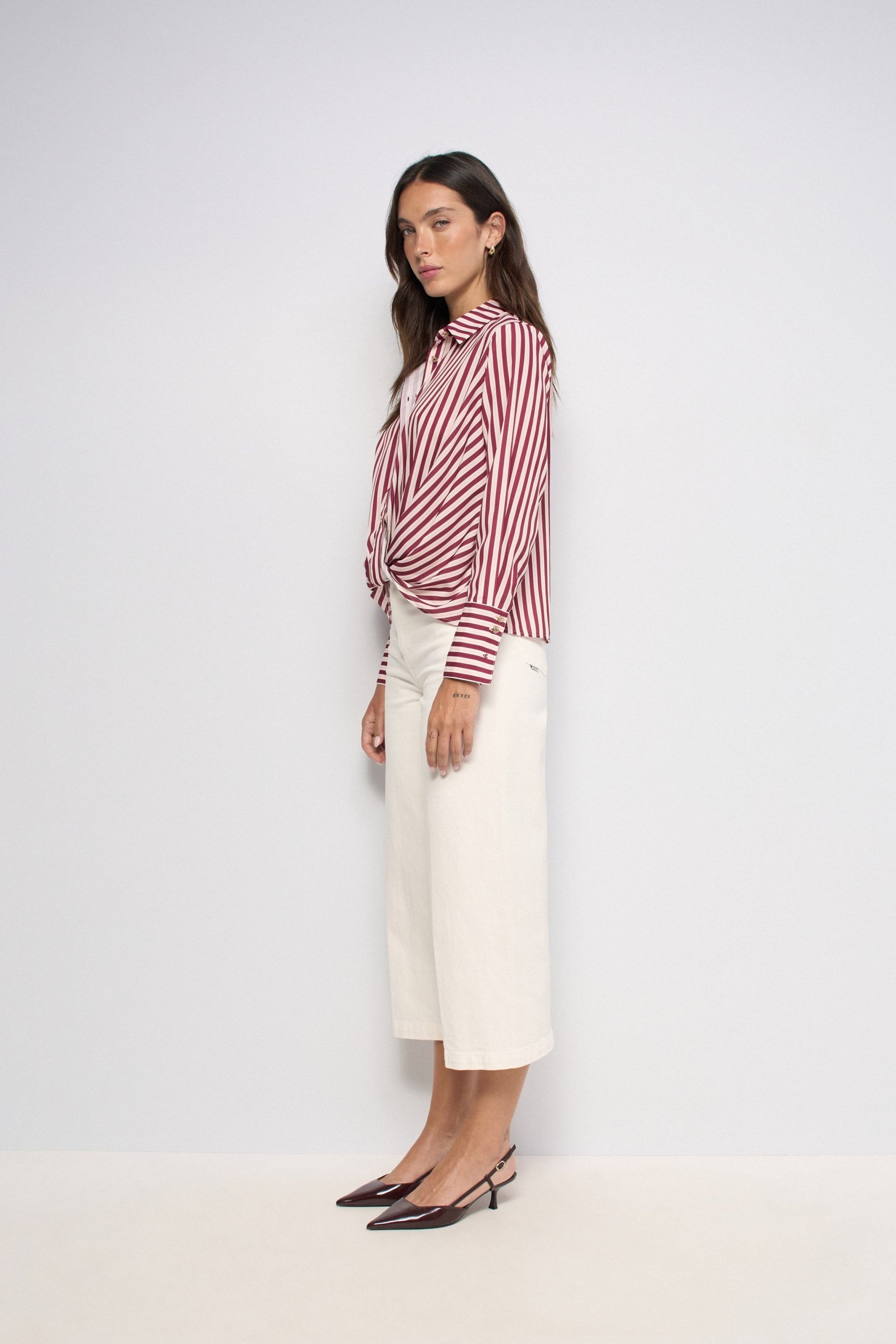 A woman stands in profile wearing a red and white vertically striped button-up shirt tied at the waist with off-white cropped trousers and dark pointed-toe flats, against a plain light gray studio background.