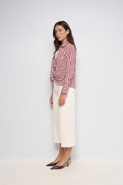 A woman stands in profile wearing a red and white vertically striped button-up shirt tied at the waist with off-white cropped trousers and dark pointed-toe flats, against a plain light gray studio background.