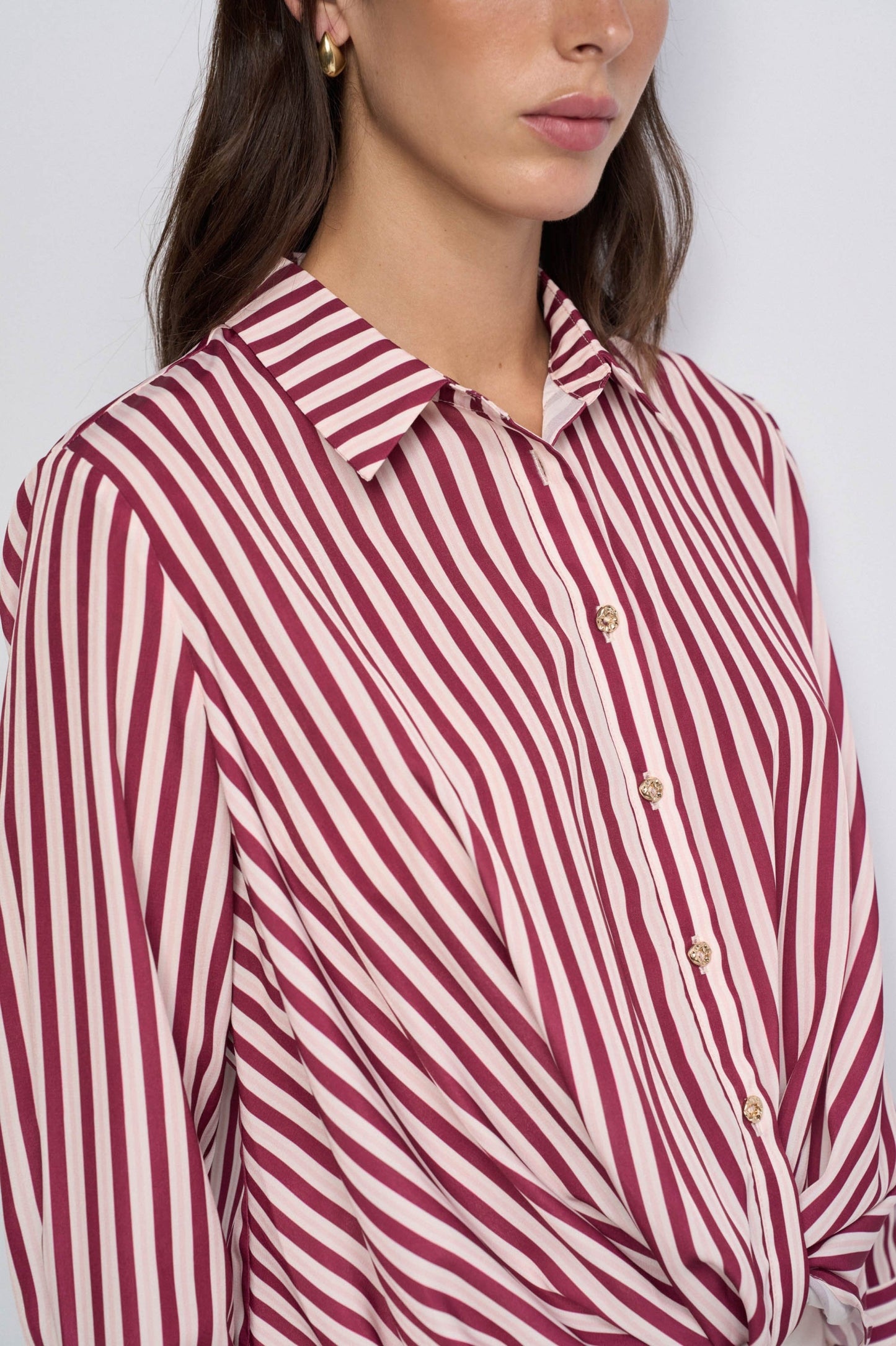 A close-up of a woman wearing a collared button-front blouse with diagonal stripes in burgundy and off-white, paired with gold-tone buttons.