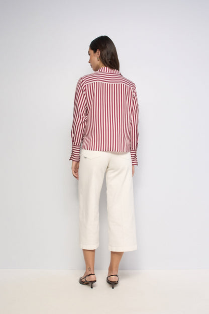 A woman is shown from the back wearing a red and white vertically striped long-sleeve button-up shirt and off-white cropped trousers, paired with black heeled sandals, against a plain light gray studio background.