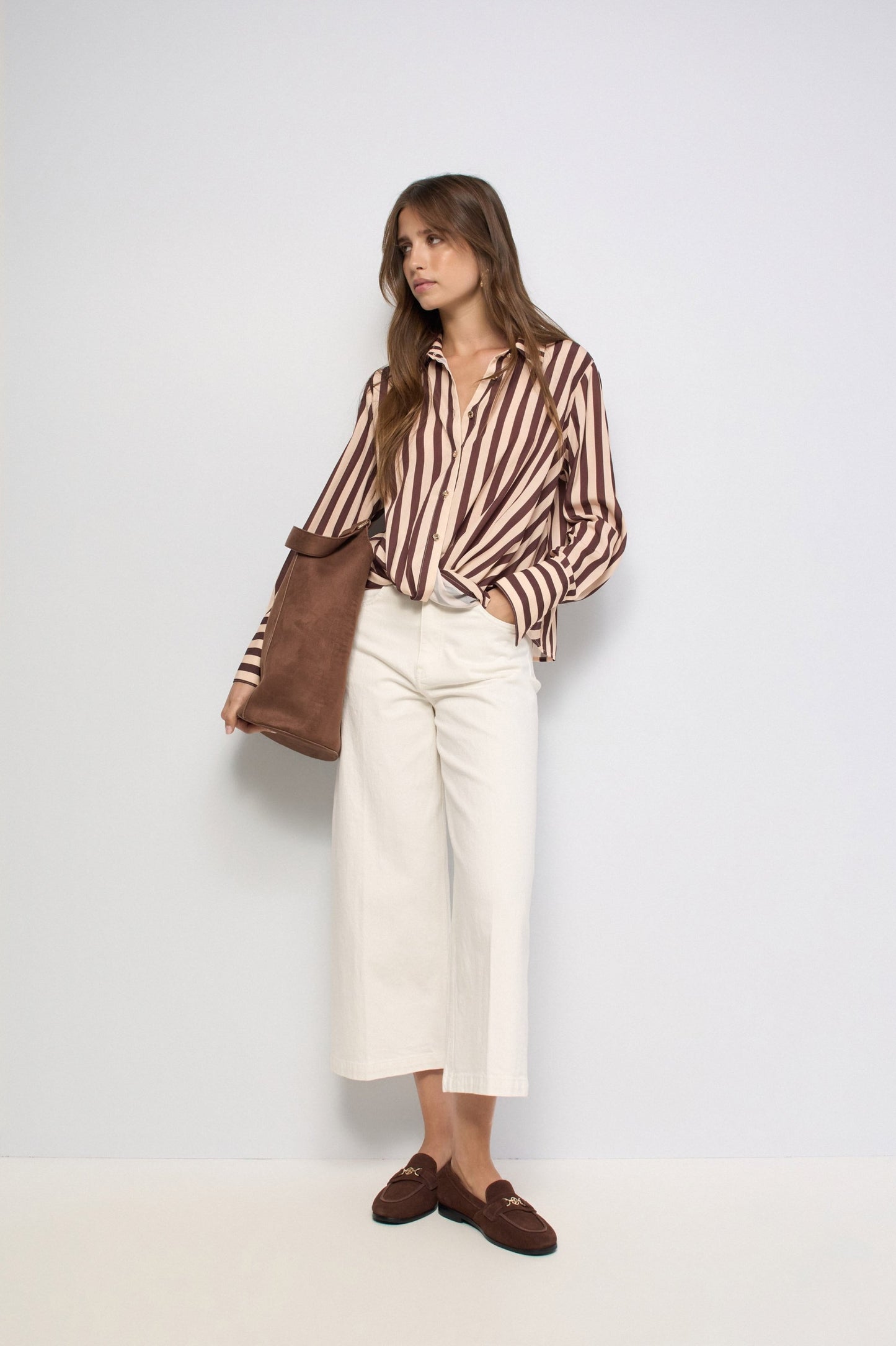 A woman stands against a plain light gray studio background wearing a brown and beige vertically striped button-up shirt, off-white cropped trousers, brown loafers, and carrying a brown tote bag.