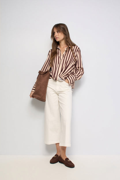 A woman stands against a plain light gray studio background wearing a brown and beige vertically striped button-up shirt, off-white cropped trousers, brown loafers, and carrying a brown tote bag.