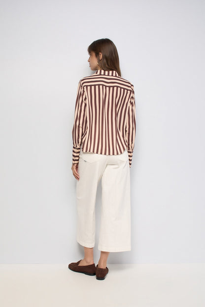 A woman is shown from the back wearing a brown and beige vertically striped long-sleeve shirt, off-white cropped trousers, and dark brown loafers, set against a plain light gray studio background.
