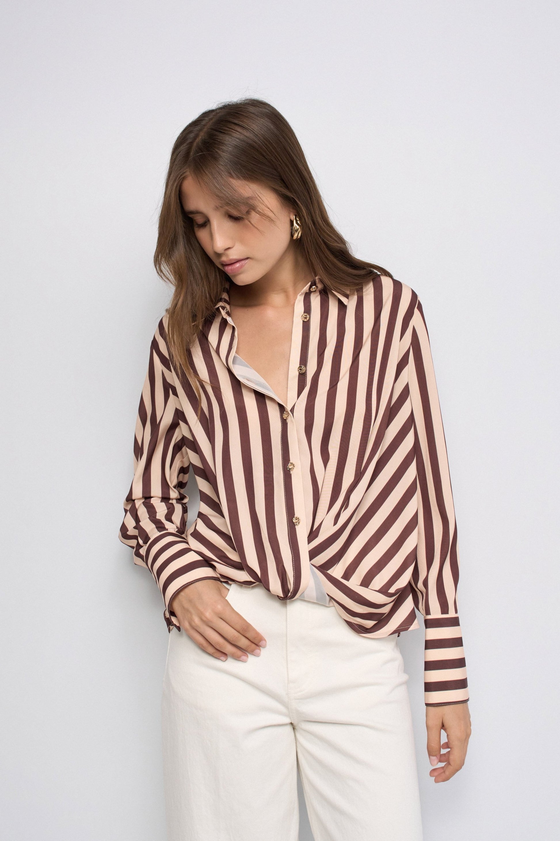 A woman wearing a brown and beige vertically striped button-up shirt tucked into white trousers, standing against a light gray studio background.
