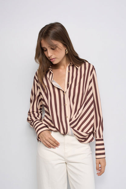 A woman wearing a brown and beige vertically striped button-up shirt tucked into white trousers, standing against a light gray studio background.