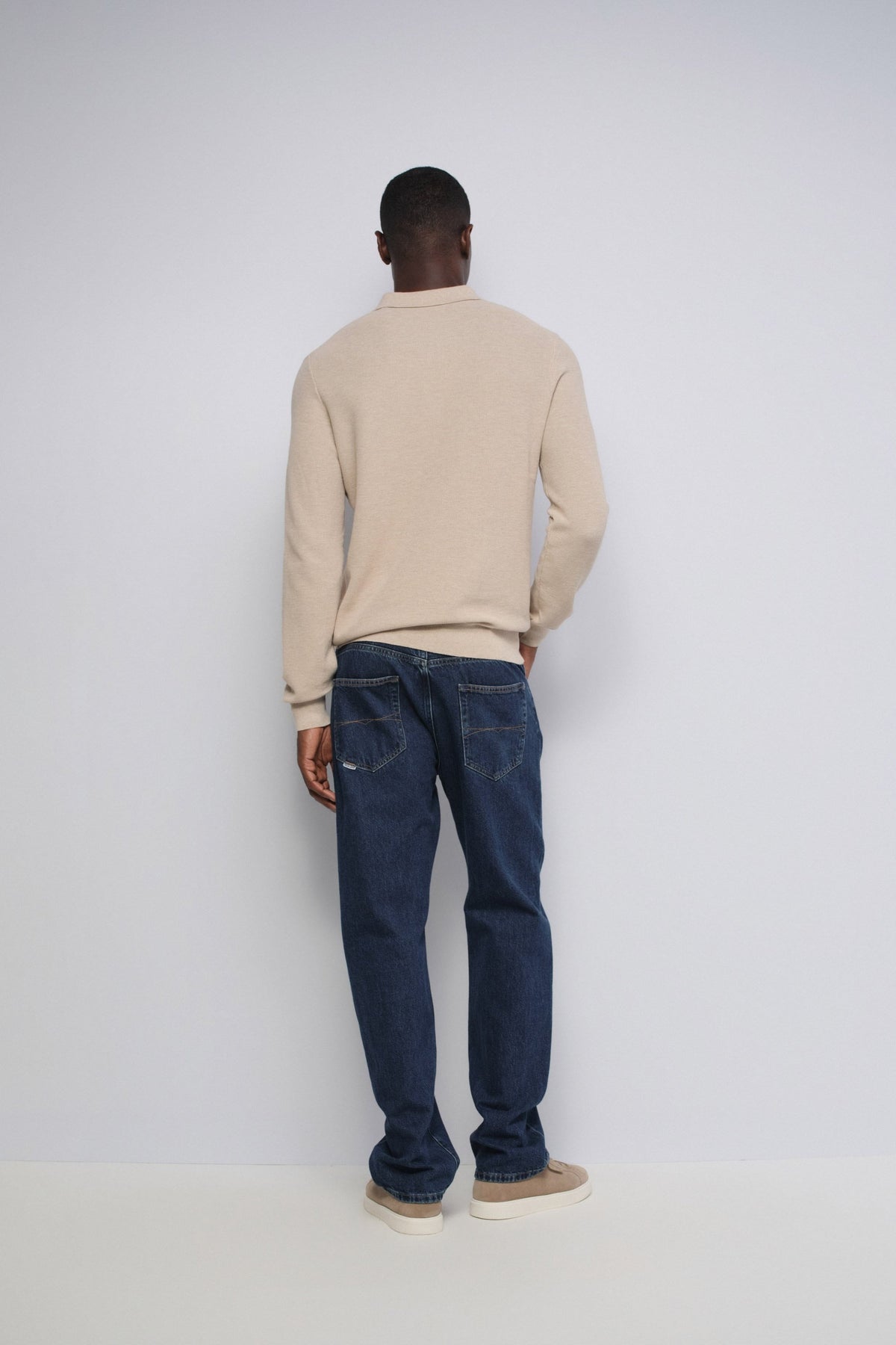 A man viewed from behind wearing a beige long-sleeve crewneck sweater and dark blue jeans, standing against a plain light gray studio background.