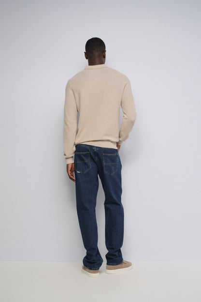 A man viewed from behind wearing a beige long-sleeve crewneck sweater and dark blue jeans, standing against a plain light gray studio background.