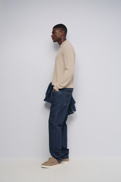 A man wearing a white Tommy Jeans t-shirt with the classic red and navy blue flag logo on the chest, paired with dark jeans and light gray sneakers, set against a plain light gray background.