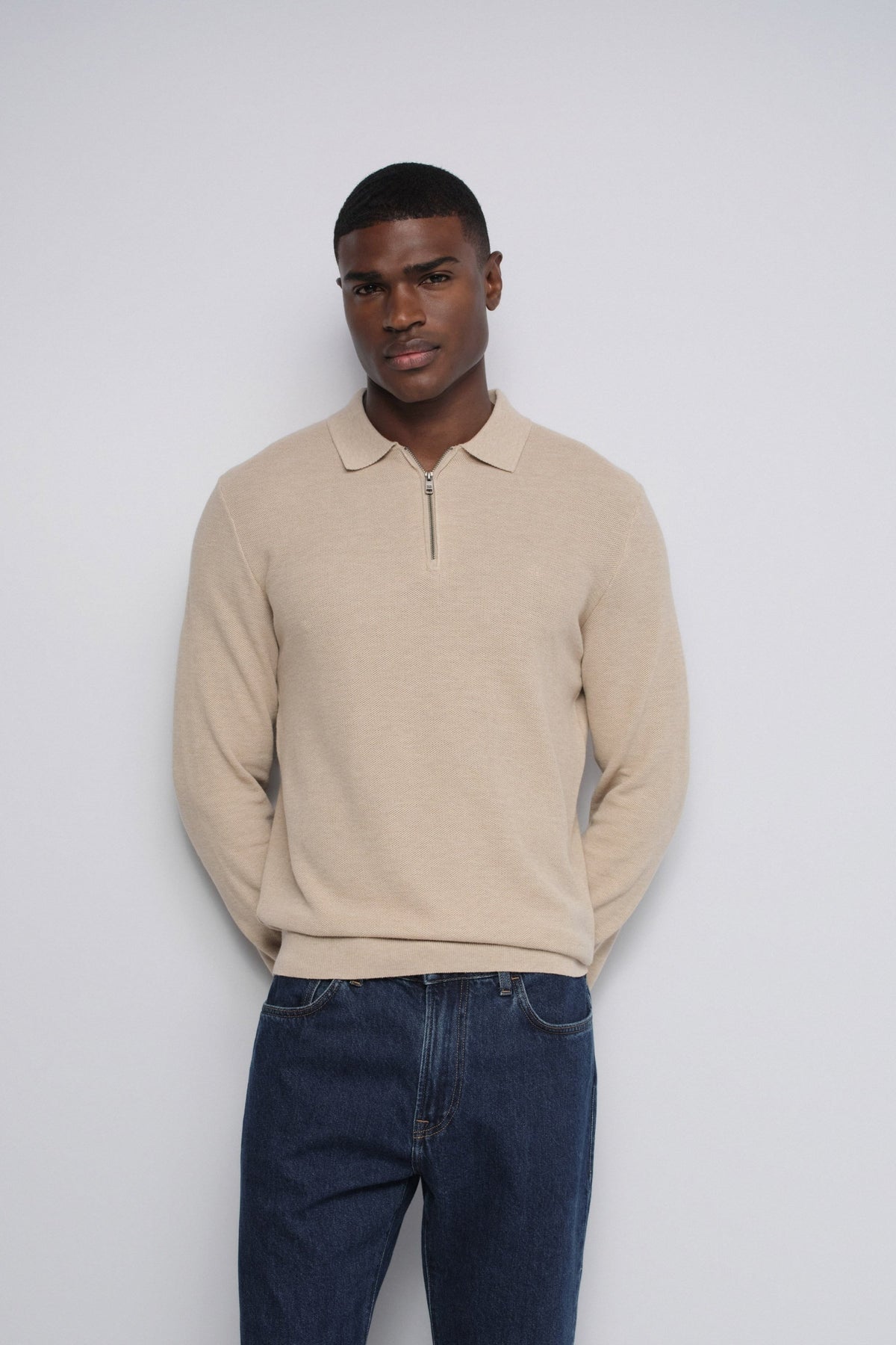 A man wearing a beige long-sleeve polo-style knit sweater with a short front zipper and dark blue jeans, standing against a plain light gray studio background.