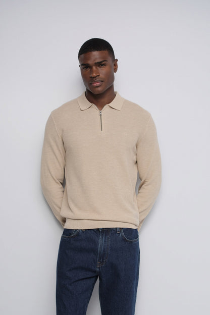 A man wearing a beige long-sleeve polo-style knit sweater with a short front zipper and dark blue jeans, standing against a plain light gray studio background.