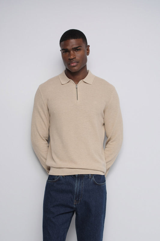 A man wearing a beige long-sleeve polo-style knit sweater with a short front zipper and dark blue jeans, standing against a plain light gray studio background.